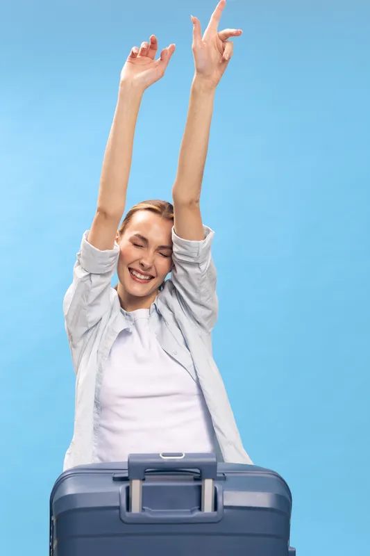 Happy young woman with suitcase stretching arms up, eyes closed and smiling on blue background. Concept of travel, vacation, freedom, relaxation, and joyful mood after journey.