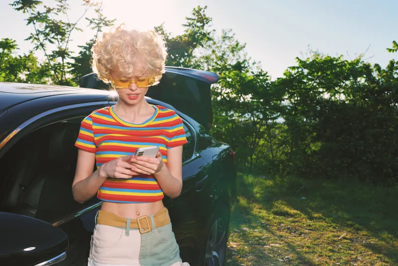 Young woman with curly blonde hair leans against a car outdoors and checks her phone, wearing a striped crop top and light shorts, conveying casual rebellious youth and carefree street vibes.