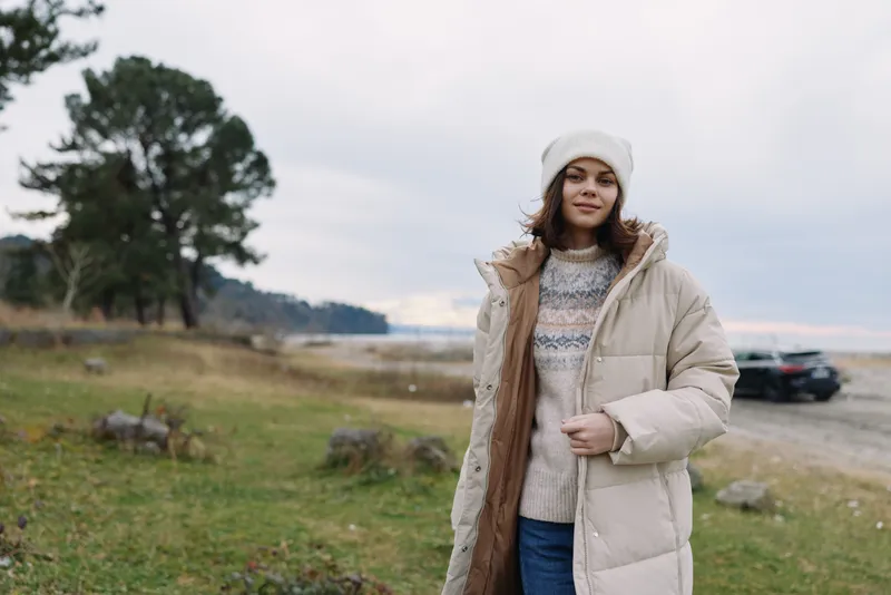 Winter fashion in an outdoor setting with a woman in a beige puffer coat and knitted sweater, standing by a coastal landscape. Calm, natural light, and relaxed mood highlight warmth and casual style.