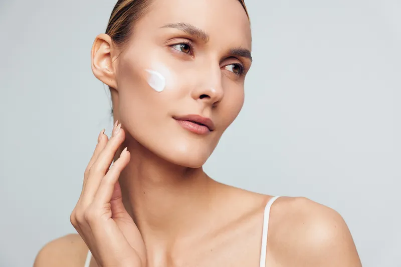Young woman with clear skin applies white face cream on her cheek while looking thoughtfully to the side against a plain light background in a skincare routine concept.
