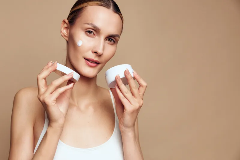 Young woman with clear skin applying moisturizing cream on face, natural beauty concept, beige background, soft lighting, skincare routine, healthy glow
