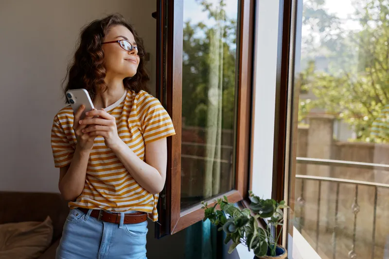 Smiling woman with curly hair standing by an open window, wearing a striped shirt and jeans. Natural daylight highlights a cozy home scene, conveying everyday lifestyle and self care.