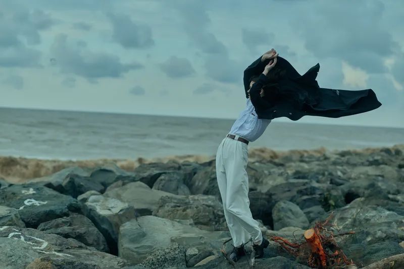 Freedom, movement, cape, wind, coastal, rocks, sea, outfit, dramatic, fashion fuse as a figure lifts a flowing cape against the shore, creating a striking, cinematic scene of resilience and