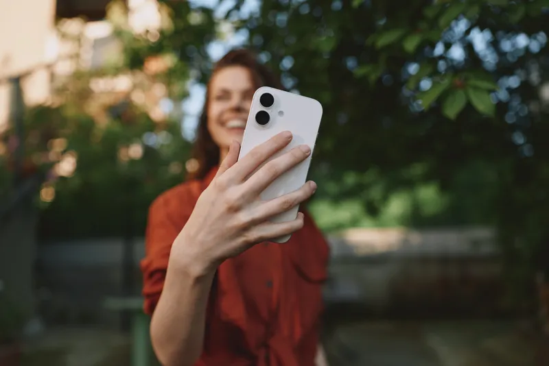 Young woman in a casual red top takes a playful selfie in a sunlit park, smiling at her smartphone and enjoying a carefree moment