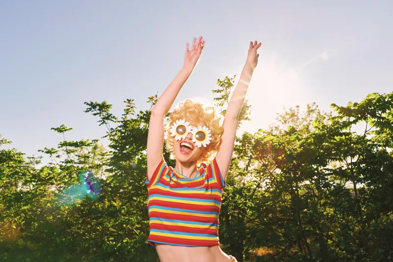 A woman outdoor summer lifestyle joy fashion retro colorful sunshine carefree stands in a sunlit park, wearing striped top and flower sunglasses, arms raised, embodying playful energy.