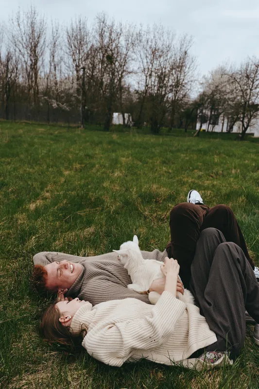 Relaxed couple lying on grass as a friendly goat kid cuddles between them, cozy sweaters, warm smiles, natural outdoor lifestyle moment, rustic park setting, calm, affectionate mood, capturing simple