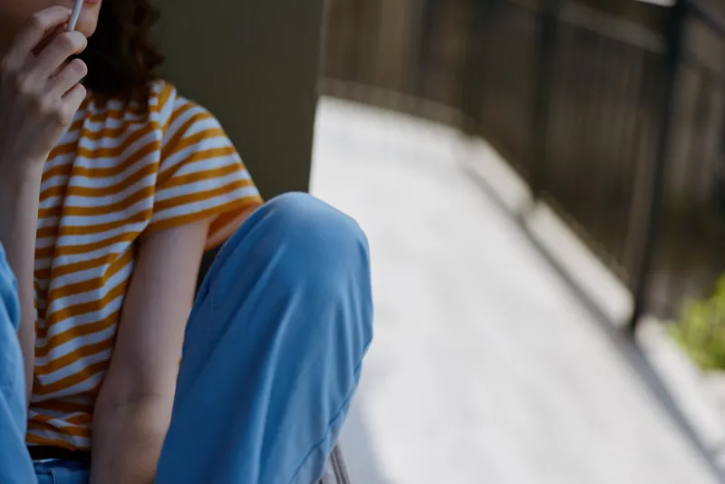A young woman sits beside a railing wearing a striped orange and white shirt and blue pants, talking on a phone with a thoughtful mood.