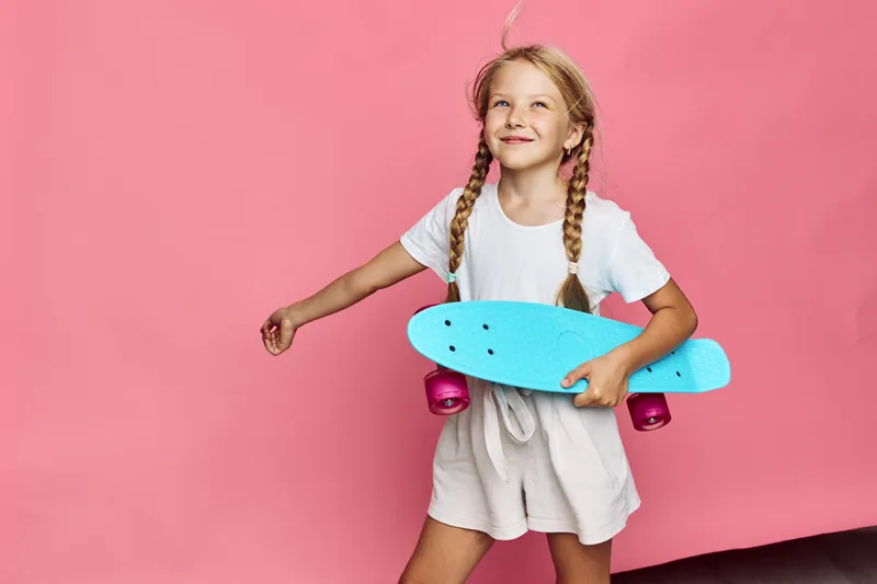 Smiling girl with braids holding a blue skateboard against pink background, wearing casual white clothes. Studio shot.