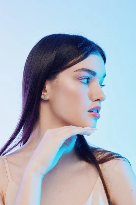Young woman with long dark hair poses in soft lighting with hand near face. Close-up profile shot on light background. Concept of beauty, fashion, and skincare.