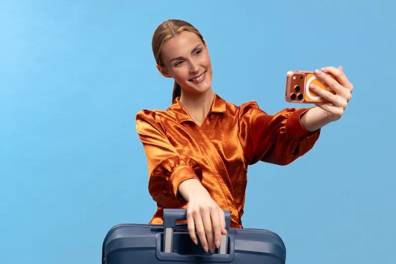 Young woman in orange satin blouse takes a selfie with smartphone while holding a blue suitcase handle on blue background. Concept of travel, vacation, and modern lifestyle.