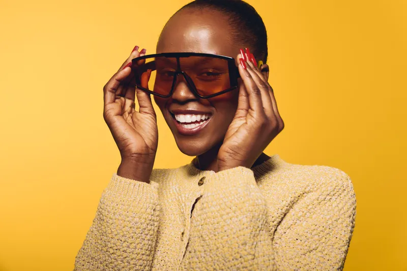 Smiling dark skinned African woman wearing oversized sunglasses and yellow knit sweater poses against bright yellow background. Concept of fashion, style, joy, and confident beauty.