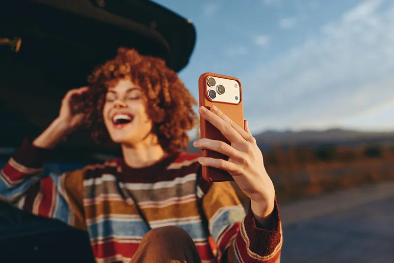 Woman smiling outdoors, laughing while taking a selfie with smartphone, curly hair in striped sweater, standing by car, bright sunny light, relaxed lifestyle, roadside landscape