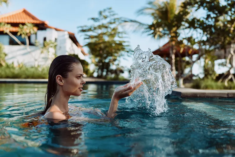 A young woman in a swimming pool splashes water with her hand during a sunny day. Tropical trees and buildings are visible in the background, lifestyle concept of relaxation.