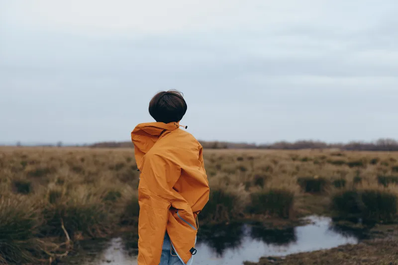 Woman outdoors lifestyle, orange jacket, walking in back, field path to river, cloudy overcast sky, natural colors, windy breeze, peaceful countryside landscape, wide horizon, calm solitude