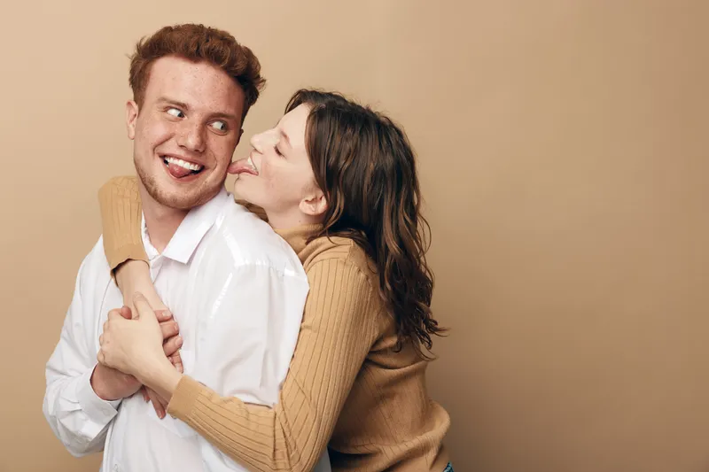 Happy young couple in casual clothes share a playful moment, woman hugging man from behind and pretending to lick his cheek. Concept of love, fun, affection, and relationship.