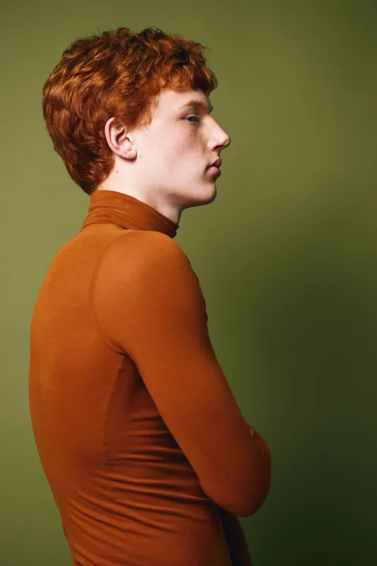 Young man with red curly hair and fair skin wearing a brown turtleneck sweater standing in profile against an olive green background in studio lighting.