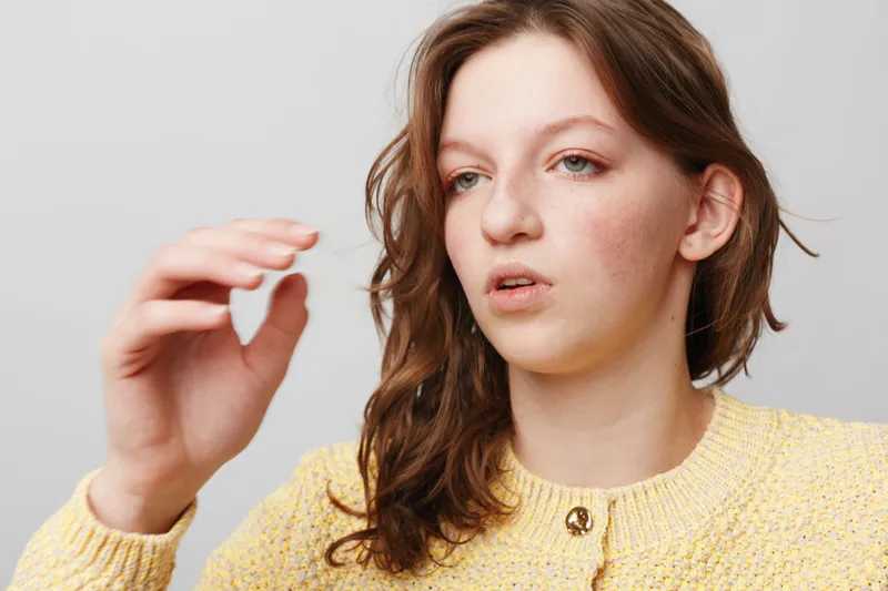 Young woman with brown hair and freckles wears yellow sweater, holding hand up with focused expression on light grey background. Concept of concentration, gesture, and casual portrait.