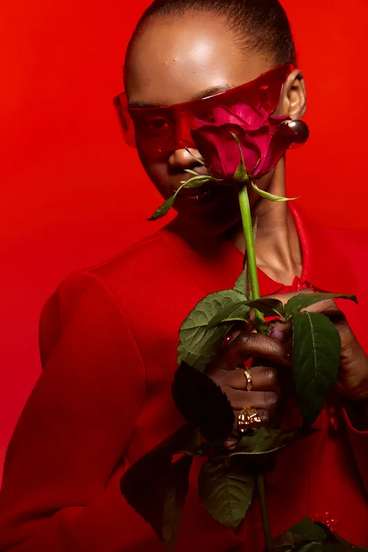 Elegant dark skinned African woman in red outfit and red sunglasses holds red rose close to face, posing on red background. Concept of fashion, beauty, love, and passion.
