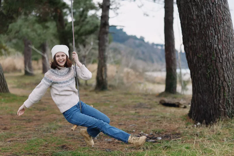 Outdoor swing scene with a playful woman in casual sweater and jeans, enjoying nature in a forest setting, capturing carefree movement and rustic charm for lifestyle stock photography