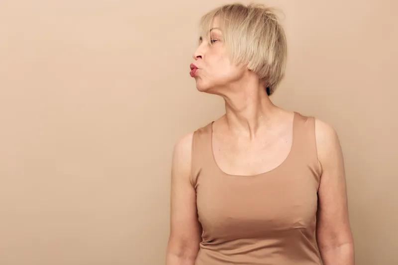 Mature woman with short blonde hair wearing beige tank top turns head to side and puckers lips as if blowing a kiss against a plain beige background. Concept of affection and playfulness.