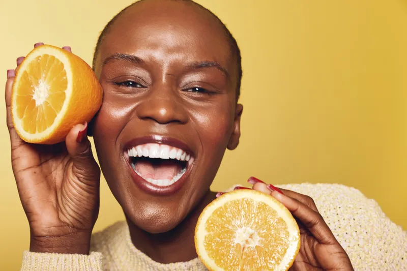 Happy black woman with dark skin holds two halves of fresh orange close to face, smiling widely. Bright yellow background and cozy sweater create warm, joyful, healthy lifestyle concept.