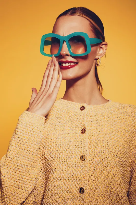 Stylish woman in oversized turquoise sunglasses and yellow knitted sweater smiles with hand near mouth on bright yellow background. Concept of fashion, accessories, and joyful mood.