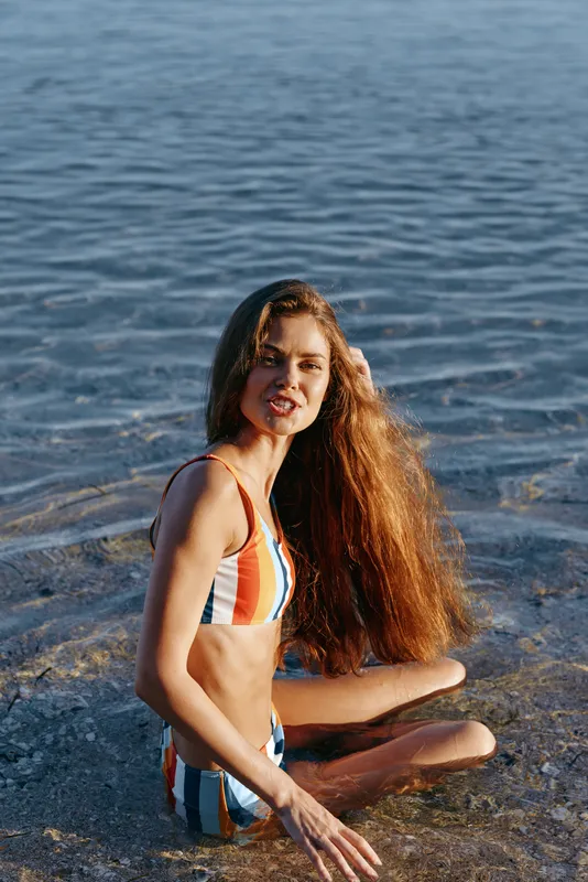 Young woman on beach at ocean waves, summer sunlight, smile in colorful swimwear, sitting on wet sand near shoreline, long red hair blowing, natural outdoor summer vacation mood, copy space
