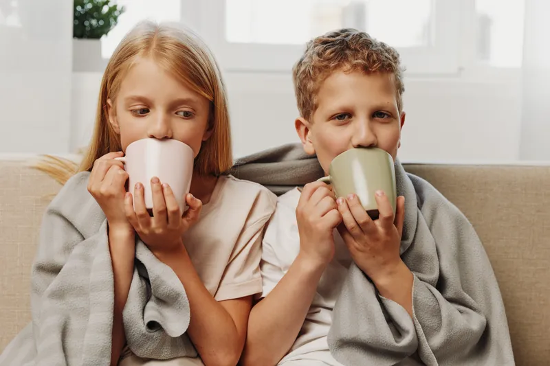 Two children wrapped in gray blankets drink hot beverages from pastel mugs at home, enjoying a cozy and calm moment together near a bright window.