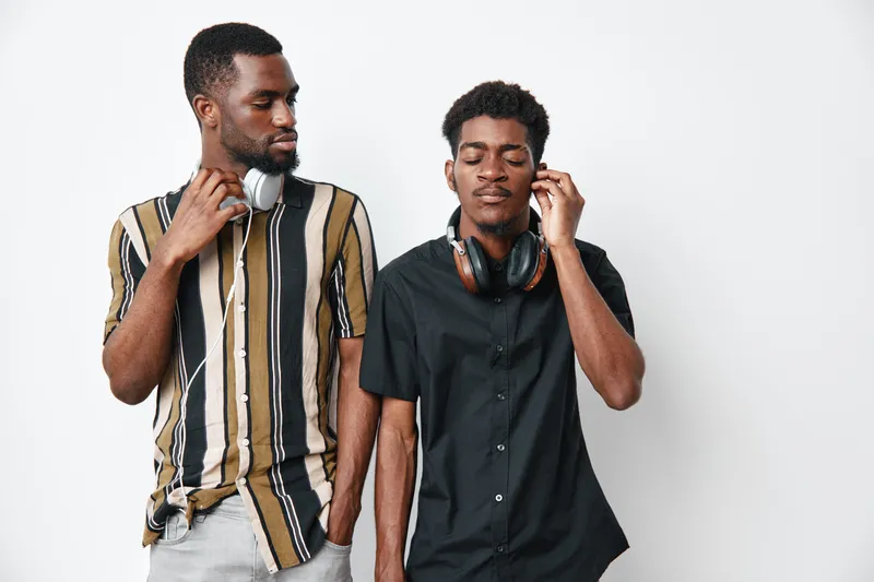 Two young African American men with dark skin wearing headphones standing together against white background. Casual style, friendship and music concept with relaxed expressions.