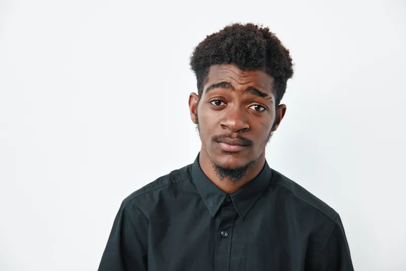 Young black man with dark skin and curly hair wearing black shirt looking at camera with neutral expression on white background. Portrait of African ethnicity male model.