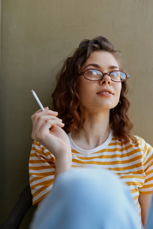 Young woman with curly hair and glasses sits indoors by a neutral wall, smoking a cigarette and enjoying a quiet, contemplative moment in a casual home setting.