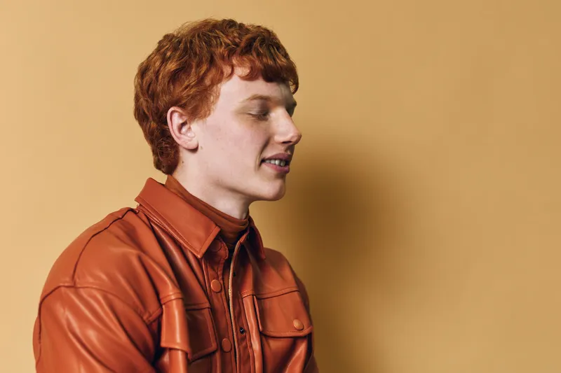 Young man with red hair wearing a brown leather jacket and shirt standing against beige background in profile with closed eyes and gentle smile, casual style portrait.