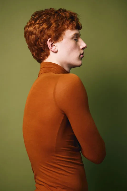 Profile of young man with red hair in orange turtleneck sweater against olive background. Side view, casual clothing and thoughtful expression with simple studio setting.