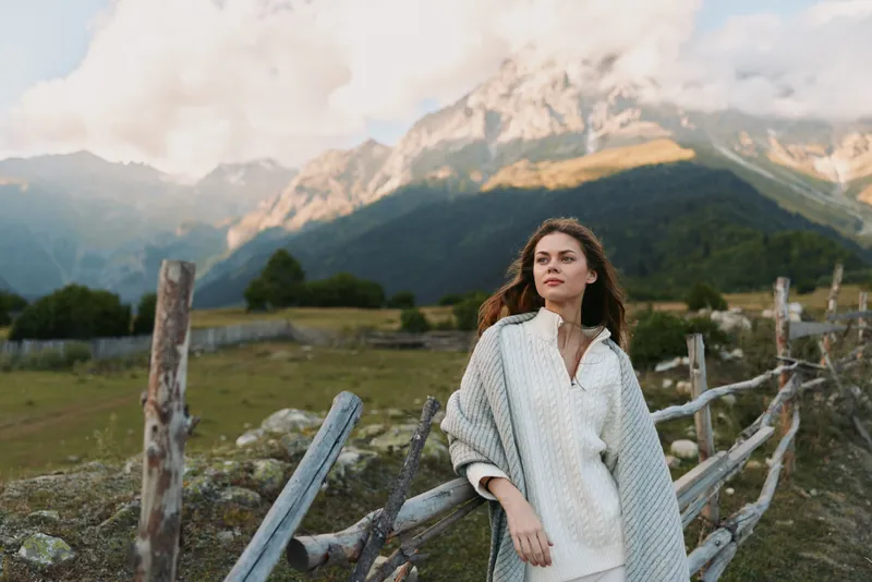 Young woman with long hair wrapped in a knitted shawl stands by a wooden fence in a mountainous landscape during a cloudy day.