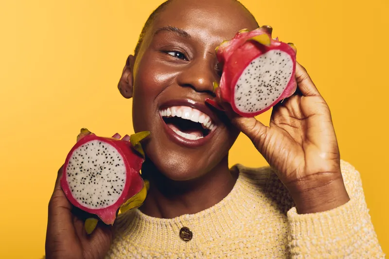 Happy black woman with dark skin smiles holding two halves of dragon fruit on yellow background. Concept of healthy eating, tropical fruit, natural lifestyle, and joyful mood.