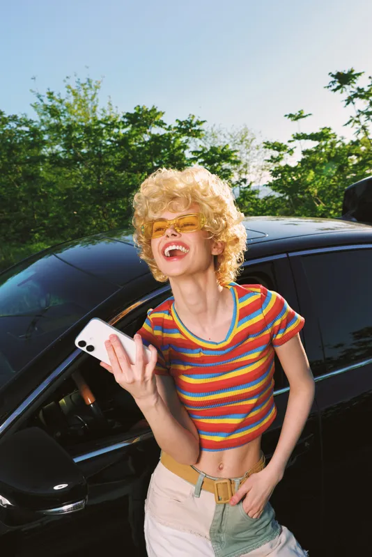 Woman lifestyle portrait with retro film color, smiling blonde in striped top beside a sleek car, holding a smartphone, carefree mood, sunlit outdoor scene, vibrant casual fashion.
