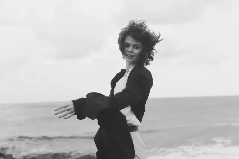 Black and white scene of a woman by the sea, wind in her hair as she drapes a jacket, capturing motion and coastal mood for fashion and lifestyle imagery