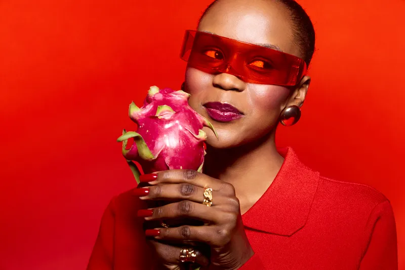 Dark skinned African woman in red outfit and red sunglasses holds a pink dragon fruit against a red background. Concept of vibrant colors, exotic fruit, fashion, and beauty.