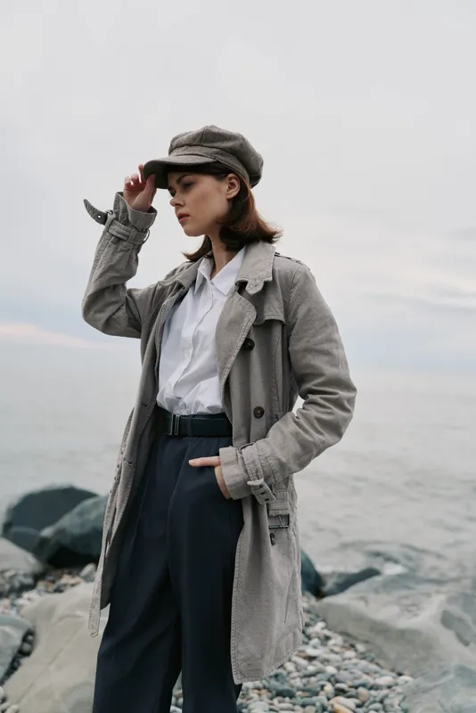 Stylish young woman in a textured coat and matching cap stands on rocky beach with ocean background. Fashion concept with casual autumn outfit and natural light mood.