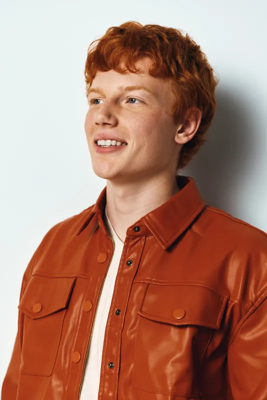 Young man with red hair wearing orange jacket smiling looking away isolated on white background. Casual style, confident expression, portrait with natural light in studio environment.