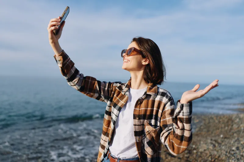 Woman on the beach taking a selfie with smartphone, wearing sunglasses and plaid shirt, casual summer vibe by the sea, confident outdoor moment with waves nearby