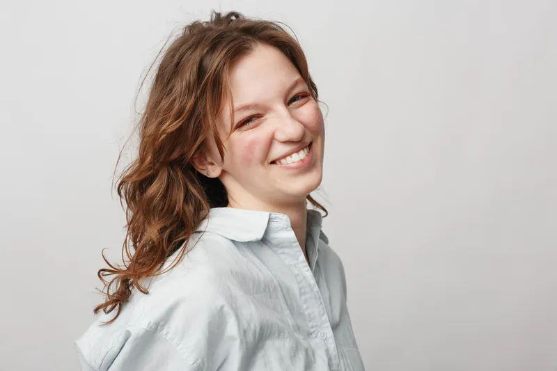 Smiling young woman with curly brown hair wearing a white shirt looks happy and confident against a plain light background. Concept of natural beauty and positive emotions.