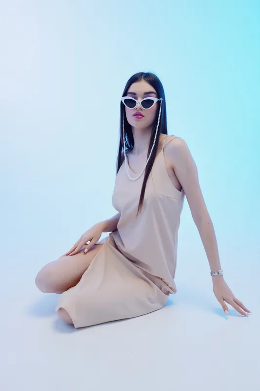 Young woman in a beige dress and white sunglasses poses seated on a white floor in a bright studio. Concept of beauty, fashion, style, and modern elegance with minimalistic background.
