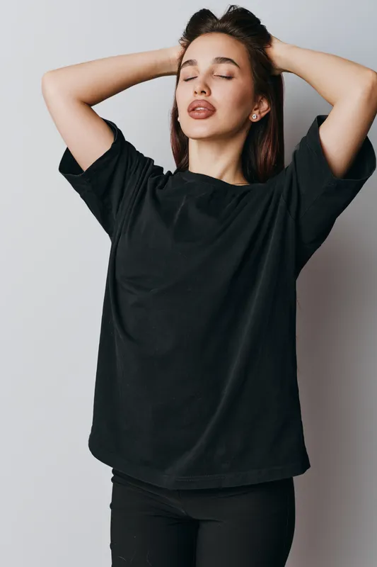 Young woman with closed eyes posing in black t shirt and black pants against white background. Casual style, relaxed expression and hands behind head for fashion portrait.