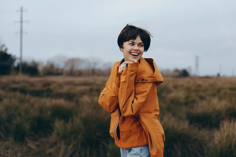 woman outdoor field smile jacket orange lifestyle nature wind portrait, smiling woman in orange jacket stands in open grass field, wind lifts hair, soft daylight for outdoor lifestyle portrait