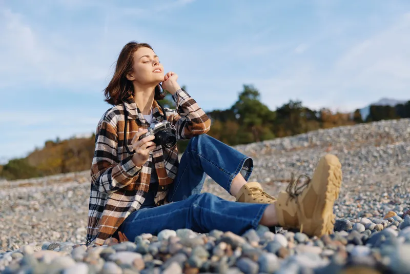 Woman sits on a pebbled shore with a camera, enjoying the outdoors in a casual plaid shirt and boots, capturing a peaceful landscape in sunlight