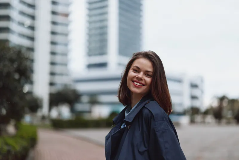 Smiling young woman with dark hair stands in a modern urban plaza, with tall buildings behind, enjoying a bright, carefree moment