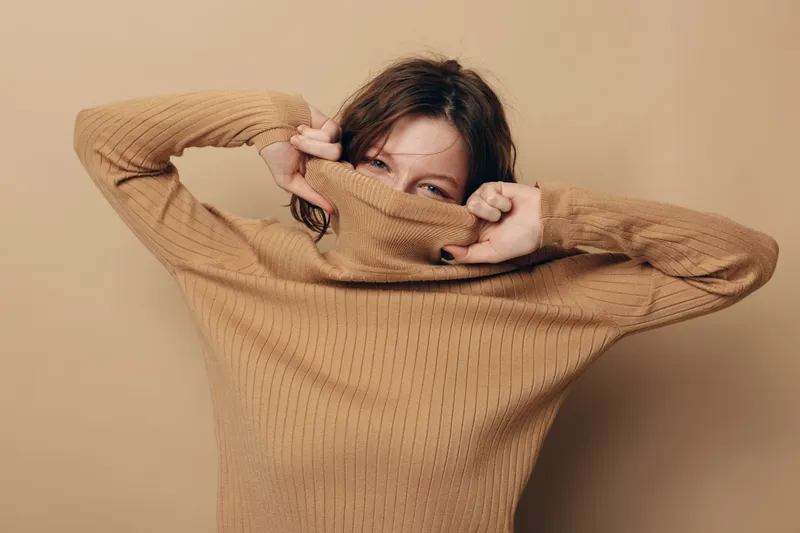 Young woman in beige knitted sweater pulls collar over face, smiling with playful eyes. Neutral background and cozy clothing create warm, casual, and relaxed mood concept.