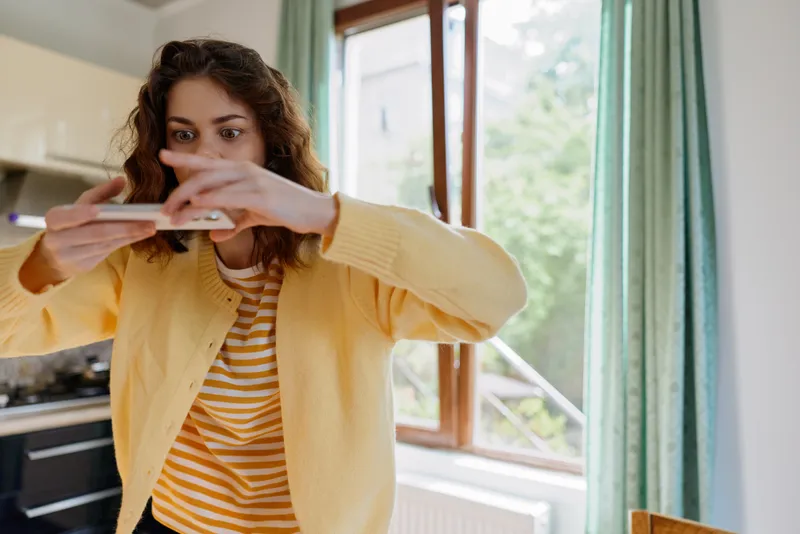 A cheerful woman in a yellow cardigan captures a casual selfie with a smartphone at home, in a bright kitchen by a window, showcasing everyday lifestyle moments.