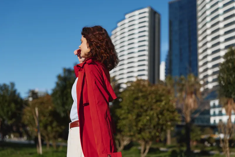 A woman in a red coat stands outdoors in a park, with tall city buildings behind her, looking to the side. Concept for urban fashion and daytime lifestyle.